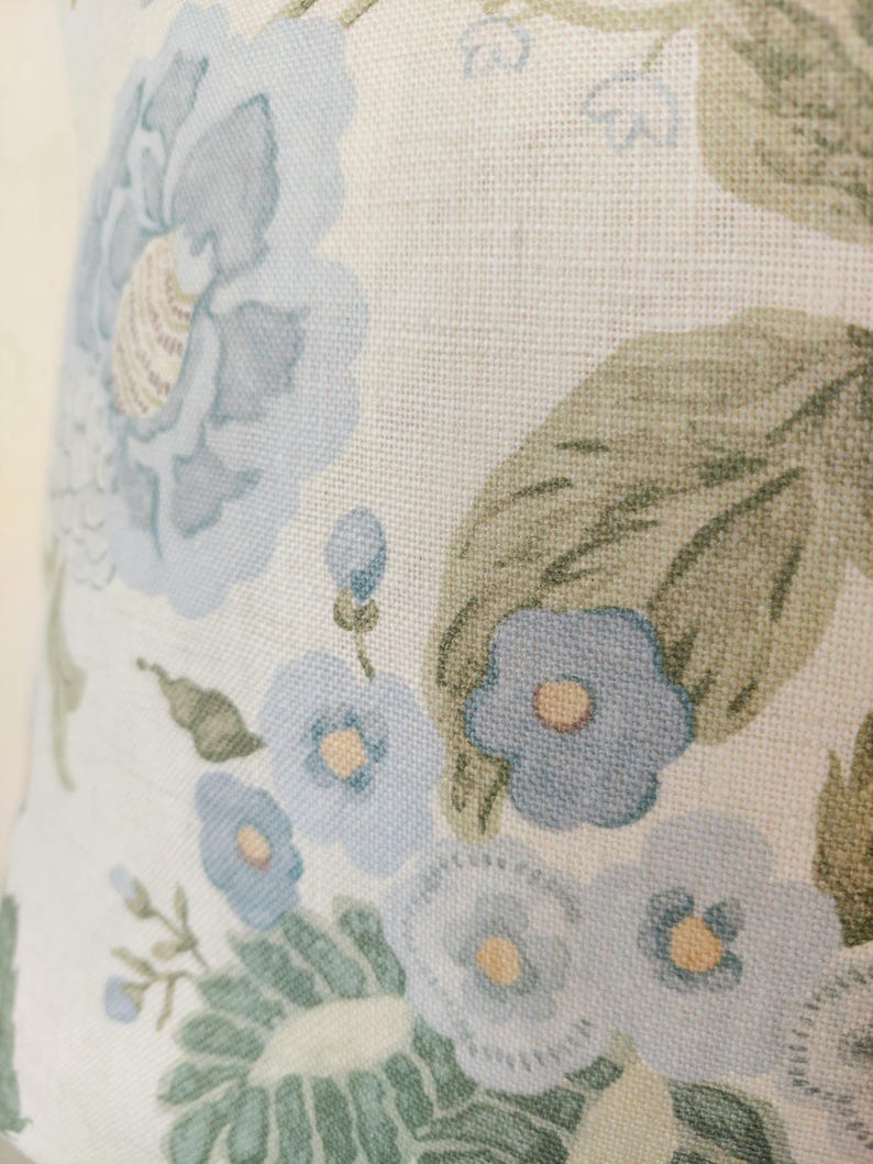 May include: Close-up of a floral pattern featuring large, light blue flowers with yellow centers and green leaves on a cream-colored background. The fabric has a textured appearance, suggesting a linen or similar material.