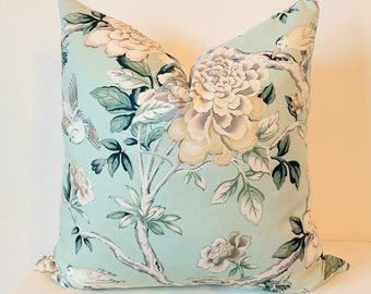 Puede incluir: Cojín decorativo cuadrado con estampado floral. El tejido azul claro presenta grandes flores color crema, hojas verdes y pequeños pájaros. El cojín está colocado sobre una superficie blanca, ideal para salón o dormitorio.