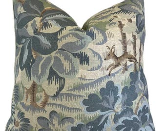 Può includere: Cuscino quadrato decorativo con un design ispirato alla natura. Il cuscino presenta un motivo di grandi elementi floreali blu e grigi, con un cervo che corre e tronchi d'albero. Lo sfondo è di colore beige chiaro.