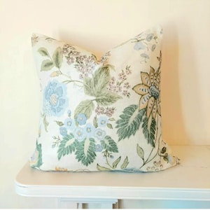 May include: A decorative square pillow with a floral pattern. The pillow features large blue, green, and tan flowers and foliage on a cream background. The pillow is resting on a white surface.