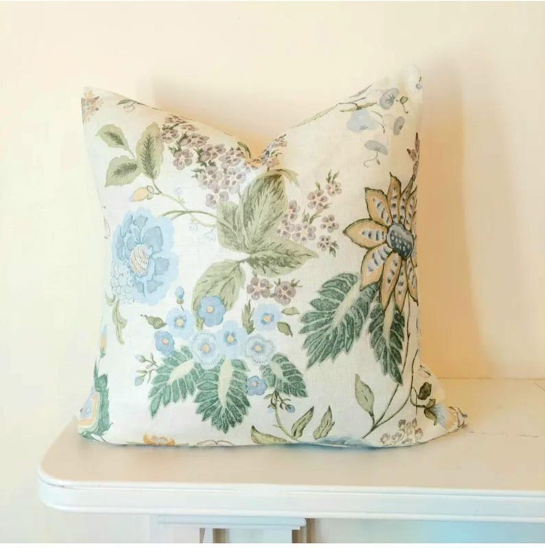 May include: A decorative square pillow with a floral pattern. The pillow features large blue, green, and tan flowers and foliage on a cream background. The pillow is resting on a white surface.