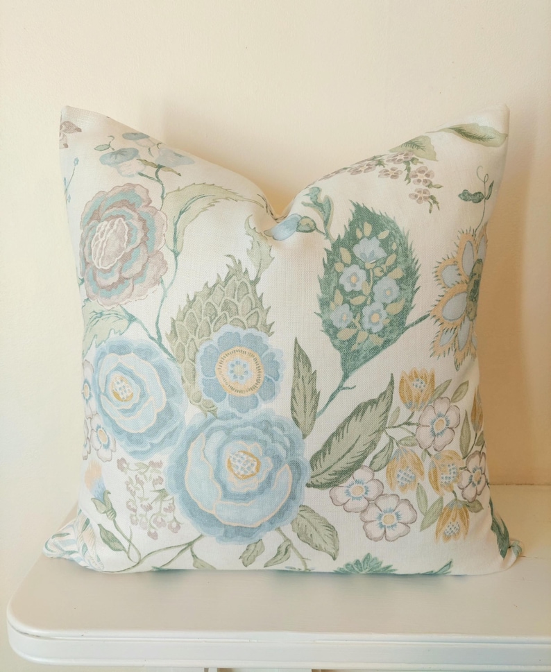 May include: A decorative pillow with a floral pattern. The pillow features large blue and pink flowers with green leaves on a cream background. The design has a vintage, botanical aesthetic, suitable for home decor.