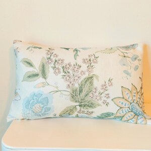 May include: Rectangular decorative pillow with a floral pattern. The pillow features a cream-colored background with light blue, green, and lavender floral designs. The pillow is placed on a white surface.