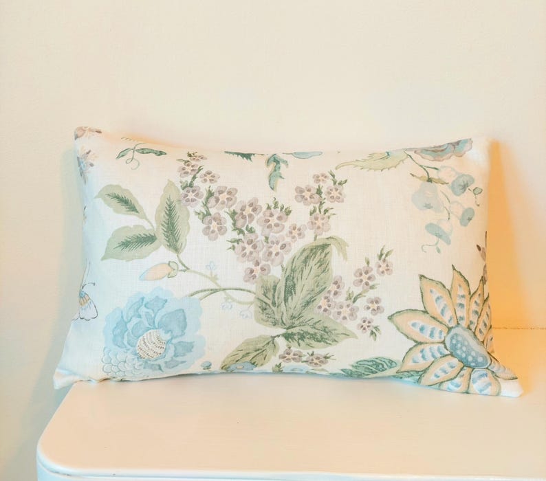 May include: Rectangular decorative pillow with a floral pattern. The pillow features a cream-colored background with light blue, green, and lavender floral designs. The pillow is placed on a white surface.