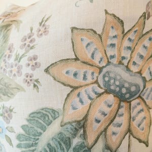 May include: Close-up of a floral pattern on a cream-colored fabric. The design features large, stylized flowers in shades of orange, blue, and green, with detailed leaves and smaller floral accents. The fabric has a textured appearance.