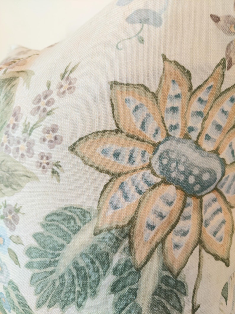 May include: Close-up of a floral pattern on a cream-colored fabric. The design features large, stylized flowers in shades of orange, blue, and green, with detailed leaves and smaller floral accents. The fabric has a textured appearance.