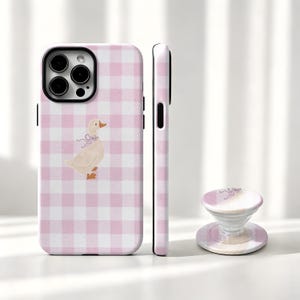 Puede incluir: Una funda para teléfono con estampado de cuadros vichy en rosa claro y blanco y un soporte retráctil a juego. La funda presenta una pequeña ilustración de un pato color crema con una cinta morada clara al cuello.