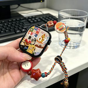 May include: A black smartwatch with a digital display showing 15:07 and a colorful collage background. The watch band is made of multi-colored braided cord decorated with various charms, including a red apple, a red star, and a hamburger icon.