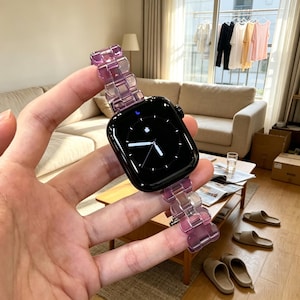 May include: A smartwatch with a black rectangular face and a translucent purple link-style watch band. The watch display shows a black background with a white analog clock face, white hands, and a small white Apple logo at the top center.