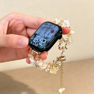 Op de afbeelding: Een zwarte smartwatch met een digitaal display dat de tijd 16:42 en een achtergrond met bamboe-thema weergeeft. De horlogeband bestaat uit goudkleurige metalen schakels afgewisseld met onregelmatige, doorschijnende gele en heldere kristalsteentjes.