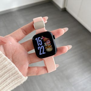 Op de afbeelding: Een smartwatch met een zwart rechthoekig scherm dat de tijd 15:22 en een stripfiguur weergeeft. Het horloge heeft een lichtroze, gestructureerde stoffen band met een zilverkleurige metalen verstelgesp voor een stevige bevestiging.