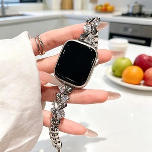 Puede incluir: Una correa de reloj inteligente en tono plateado que presenta una serie de eslabones metálicos interconectados, cada uno con un cristal transparente facetado en forma de lágrima. La correa está unida a un reloj inteligente rectangular.