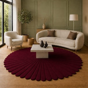 Puede incluir: Un salón con una alfombra burdeos en forma de pétalo. Un sofá beige con cojines marrones se encuentra frente a una pared verde oliva. También hay un sillón blanco y una mesa de centro cuadrada con jarrones negros.