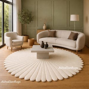 May include: A cream-colored, round rug with a petal-like design dominates the foreground of a living room scene. A beige sofa, armchair, and square coffee table complement the rug. The room features a neutral color palette with wooden floors and a large window.