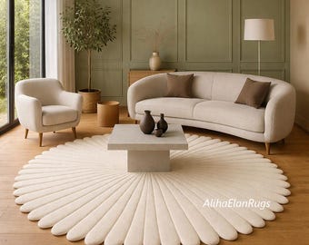 May include: A cream-colored, round rug with a petal-like design dominates the foreground of a living room scene. A beige sofa, armchair, and square coffee table complement the rug. The room features a neutral color palette with wooden floors and a large window.