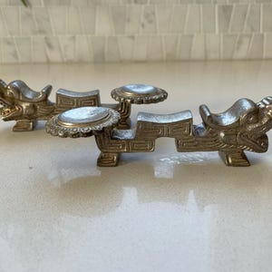 Könnte beinhalten: Zwei silberfarbene Stäbchenhalter in Drachenform. Jeder Halter hat einen Drachenkopf an einem Ende und eine kleine Plattform für Essstäbchen. Die Halter haben ein detailliertes, verziertes Design und sind aus Metall.
