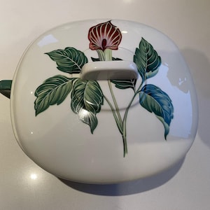 May include: White ceramic casserole dish with a lid featuring a floral design. The lid has a handle and a green accent on the side. The floral design includes a red and white flower with green leaves.