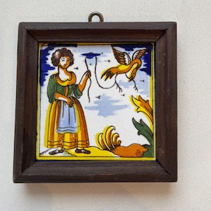 May include: A framed ceramic tile with a folk art design. The tile features a woman in a green and yellow dress, a yellow bird, and floral elements. The frame is dark brown and square, with a metal loop for hanging.