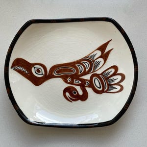 May include: A white ceramic dish with a brown bird design. The dish has a black rim and a unique shape. The bird design is stylized with intricate details, and the dish has a crackled surface.