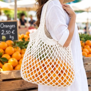 Puede incluir: Una bolsa de mercado de ganchillo color crema con un patrón de malla en forma de diamante y dos asas integradas. La bolsa está llena de cítricos de color naranja brillante, mostrando la construcción de tejido abierto y el color vibrante del contenido.