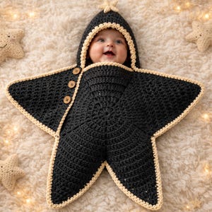 Puede incluir: Una manta de arrullo para bebé tejida a ganchillo en color negro con forma de estrella y capucha, que presenta un borde de color crema y tres botones de madera en el lateral. Incluye una pequeña estrella tejida en la parte superior.