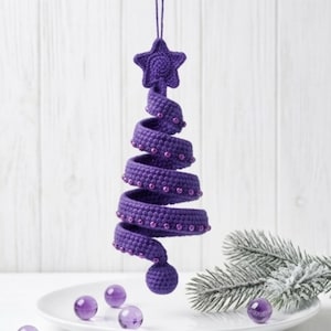 Puede incluir: Un adorno en espiral de ganchillo morado con forma de árbol, rematado con una estrella de cinco puntas. La espiral está decorada con pequeñas cuentas moradas a juego a lo largo del borde, y una pequeña esfera morada cuelga de la punta.