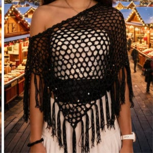 May include: A black crochet poncho featuring an open net pattern and long fringe trim along the bottom edge. The garment is designed with an asymmetrical one-shoulder neckline and is worn over a plain white top, creating a layered appearance.