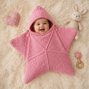 Puede incluir: Un bebé envuelto en una manta de arrullo tejida en forma de estrella rosa con capucha. La manta presenta un patrón de punto trenzado texturizado y dos cremalleras laterales. Un pequeño cojín rosa en forma de corazón está cerca.