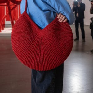 Puede incluir: Un bolso de hombro circular grande de ganchillo en color rojo intenso, con un patrón de punto acanalado texturizado que irradia desde el centro. El bolso tiene una sola correa integrada y una silueta ancha y redondeada de hilo grueso.