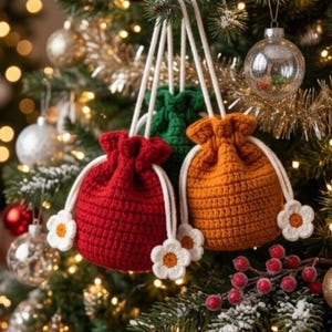 May include: Three small crochet drawstring pouches in red, green, and orange, each featuring white cord drawstrings finished with small white and yellow flower-shaped accents. The pouches are suspended by their cords, showcasing a textured stitch.