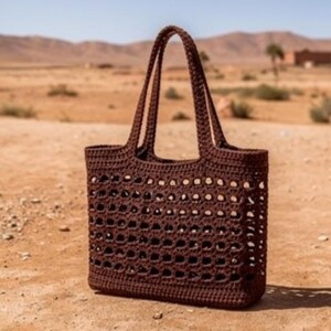Puede incluir: Un bolso tote de ganchillo en color marrón oscuro con base rectangular y dos asas largas para el hombro. El cuerpo del bolso presenta un patrón de rejilla calado repetitivo, mientras que el borde superior y la base tienen un tejido denso.