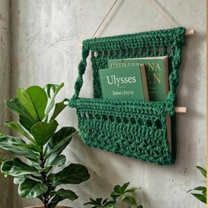 Puede incluir: Un organizador de pared de ganchillo en color verde oscuro suspendido por un palo de madera y un cordón fino. El bolsillo contiene dos libros verdes, uno de ellos titulado Ulysses de James Joyce, con texto visible en la portada en letras blancas.