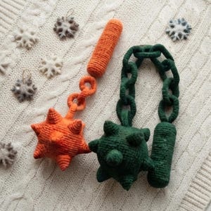 May include: Two crocheted flail toys, one orange and one dark green, each featuring a spiked ball head, a short handle, and a flexible chain link connector. Several small, felted white and gray snowflake ornaments are scattered nearby.