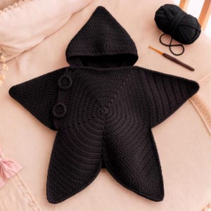 May include: A black crocheted star-shaped hooded garment featuring a pointed hood and two circular button-like closures on one side. The piece is crafted with a consistent stitch pattern and is accompanied by a ball of black yarn and a crochet hook.