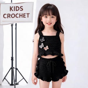 Puede incluir: Un conjunto de dos piezas de ganchillo negro que consta de un top corto sin mangas con tres pequeñas aplicaciones de mariposas y pantalones cortos a juego con cintura de cordón y bordes con volantes. Un letrero dice KIDS CROCHET.