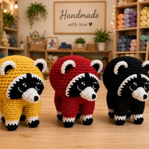Puede incluir: Tres peluches de mapache tejidos a ganchillo en fila. El de la izquierda es amarillo, el del centro es rojo y el de la derecha es negro. Cada mapache tiene una mancha blanca en la cara, antifaz negro, grandes ojos negros y patas blancas.