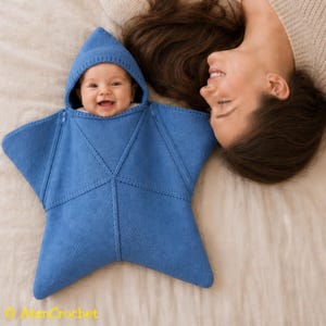 May include: A blue knitted baby swaddle blanket shaped like a five-pointed star with a pointed hood. The garment features a central seam design and zips along the upper edges. The text AtenCrochet is visible in yellow in the bottom left corner.