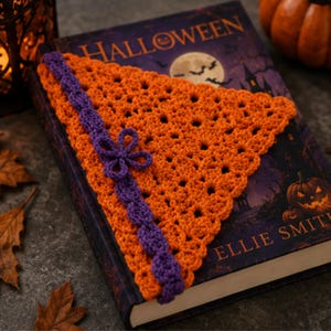 Puede incluir: Un marcapáginas triangular de ganchillo naranja con un patrón de punto calado y un borde de ganchillo morado con un pequeño adorno de flor morada. El marcapáginas está colocado sobre un libro titulado Halloween con una luna.