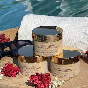 Op de afbeelding: Drie doorzichtige plastic potten met gouden deksels met HumbleGlow Collections NurseCrafted Brightening Body Scrub. De etiketten vermelden Turmeric, Kojic Acid en Niacinamide, met een nettogewicht van 113 g per pot op een houten dienblad.