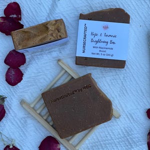 Op de afbeelding: Twee rechthoekige bruine zeepblokken met een textuur. Eén blok heeft een wit etiket met de tekst "NURSECRAFTED Kojic & Turmeric Brightening Bar With Niacinamide Boost Net Wt. 5 oz (141 g)". Het andere blok is voorzien van de inscriptie "NurseCrafted by HQC".