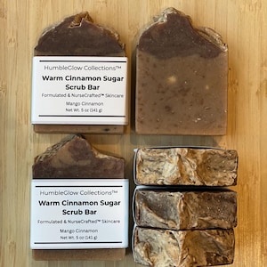 May include: Four bars of handmade soap are displayed on a wooden surface. Two bars are labelled "Warm Cinnamon Sugar Scrub Bar" with "Mango Cinnamon" and "Net Wt. 141 g". The soap has a brown and tan marbled appearance.