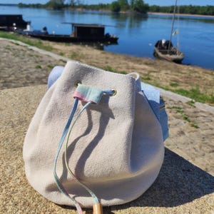 Pode incluir: Uma bolsa bege com cordão e um detalhe em cores pastel e cordões. A bolsa tem ilhós dourados e contas de madeira nos cordões. O fundo mostra um rio com barcos e um céu azul claro.