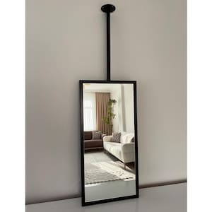 Pode incluir: Um espelho retangular com moldura preta suspenso do teto por uma haste preta. O espelho reflete uma sala de estar com um sofá branco, um sofá marrom e um tapete estampado. O espelho é um item de decoração para casa.