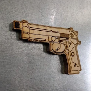 May include: A wooden cutout of a handgun, featuring detailed laser-cut lines. The gun is light brown and appears to be made of plywood. The image is taken from a slightly angled perspective, showing the side of the firearm.