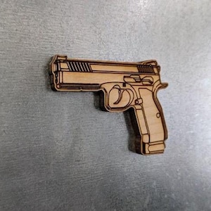 Puede incluir: Un recorte de madera de una pistola, con contornos detallados de la corredera, el gatillo y la empuñadura. La veta de la madera de color marrón claro es visible, sobre un fondo gris. El recorte es una representación detallada de un arma de fuego.