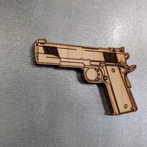 May include: A wooden pistol cutout with detailed laser-cut lines. The light brown wood has darker brown accents. The gun is a detailed replica of a handgun. The cutout is likely a decorative item or craft supply.