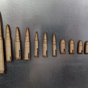 Puede incluir: Una colección de réplicas de balas de madera de varios tamaños, dispuestas de mayor a menor. Las balas son de color marrón claro y tienen diseños detallados, incluyendo la punta y la carcasa. El fondo es una superficie gris metálico.