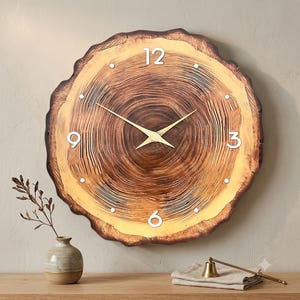 May include: A round wall clock featuring a natural wood grain pattern that mimics a tree cross-section. The clock face displays white numerals at the 12, 3, 6, and 9 o'clock positions, with simple white dots for other hours and thin gold-toned hands.