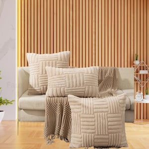 May include: Beige decorative throw pillows with a geometric pattern are arranged on a beige sofa, along with a matching throw blanket. The pillows have a textured, raised design. The background features a wooden slatted wall.