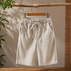 May include: Beige shorts featuring an elasticized waistband with a matching drawstring tie at the center. These knee-length shorts are constructed from a textured fabric and include two side pockets, providing a simple and functional design.